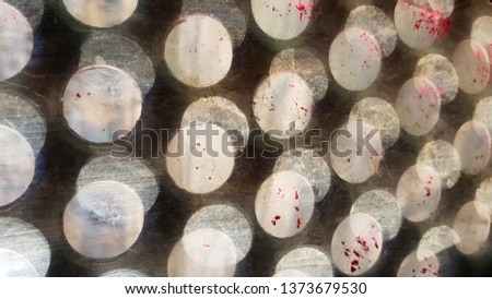 Blurred dotted multilayered glass background. Grunge textures of dirty glass surface closeup through round hole lattice window. Abstract fuzzy background with dots of lights and stains of red paint 
