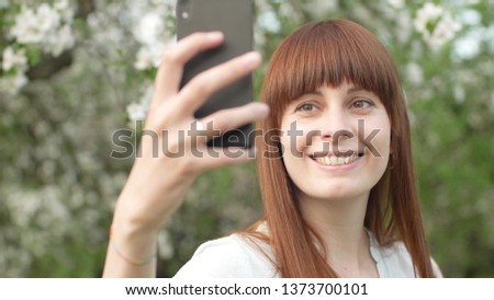 A girl makes selfie in the garden. An attractive red-haired woman smiles making selfi using a mobile phone in a cherry orchard. The concept of using gadgets for a healthy lifestyle.