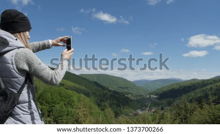 A woman is taking pictures on the phone against the background of big mountains and the green mountain river. selfie or self-portrait on a smartphone. Enjoys adventure and travel concept