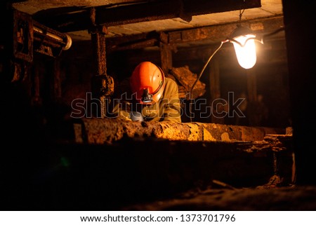 A young guy in a protective suit and helmet sits in a tunnel with a burning scrapbook. Miner in mine Royalty-Free Stock Photo #1373701796