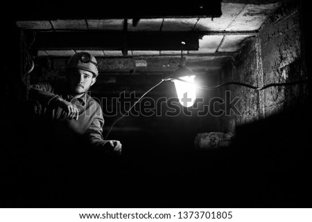 A young guy in a protective suit and helmet sits in a tunnel with a burning scrapbook. Miner in mine Royalty-Free Stock Photo #1373701805