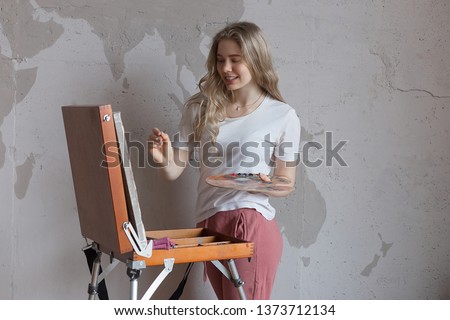 Young smiling pretty blonde girl with brush and palette standing near easel drawing picture. Art, creativity, hobby concept, painting process.
