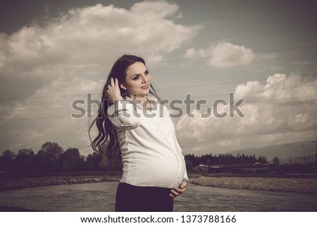 Pregnant woman posing at park near to river. Lovely hipster woman standing in the park cloudy weather
