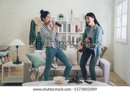 Young happy girl best friends having party one playing guitar and another singing by holding beer bottle as microphone. two asian women dancing together. Friendship leisure rest home enjoy concept Royalty-Free Stock Photo #1373802089