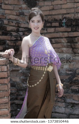 Thai Woman, wear the hair in a bun, posing and wearing in Thai Ayutthaya Traditional costume with purple clothes with a blurred wall built in stones and red brick background. Historical and fashion.