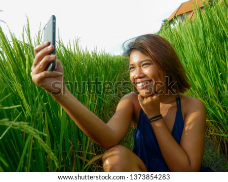 young happy and exotic islander Asian girl from Indonesia taking selfie self portrait photo with mobile phone smiling cheerful and excited posing rice field nature background