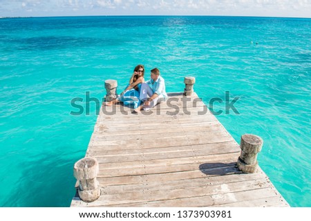 Couple having good time at the sea. They are sitting at the wooden pier, hugging and smiling. Turquoise water is around them. Drone view picture.