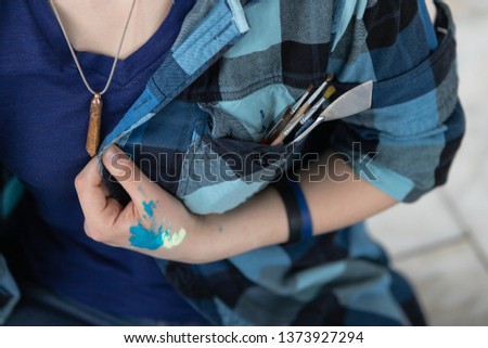 Paint brushes and palette knife placed in the pocket of a checkered shirt. The girl with paints on a hand holds a shirt. Oil painting tools.