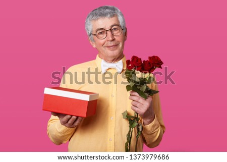 Close up portrait of good looking eldery man in spectacles, dressed in yellow shirt in one tone and white bowtie, carries red box with present and red flowers as present for birthday. People concept.