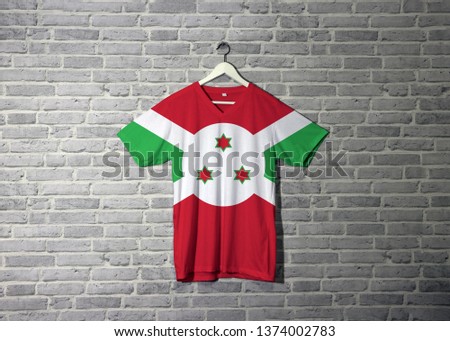 Burundi flag on shirt and hanging on the wall with brick pattern wallpaper. A white diagonal cross divided into four panels of red and green and three star in the white disk in the center. 