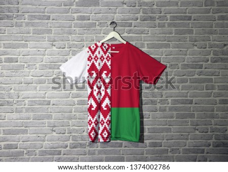 Belarus flag on shirt and hanging on the wall with brick pattern wallpaper, a horizontal bicolor of red over green in a 2:1 ratio, with a red ornamental pattern on a white.