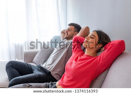 Couple relaxing on a sofa at home and looking at TV at the living room. Side view of a happy couple breathing and resting lying in a couch at home with a window in the background