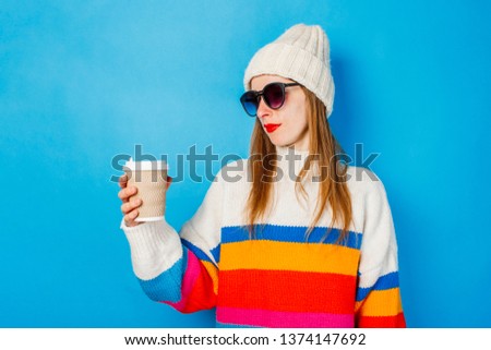 A young girl in glasses and a hat is holding a paper cup with coffee or tea and looking at it against a blue background. Concept coffee shop, winter, hipster.