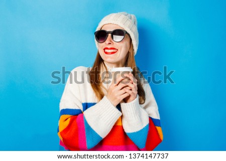 A young girl in glasses and a hat is smiling and holding a paper cup with tea or coffee against a blue background. Concept coffee shop, winter, hipster.