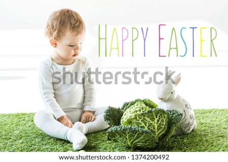 child sitting near savoy cabbage and decorative rabbit on green grass with happy Easter lettering