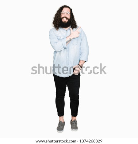 Young hipster man with long hair and beard wearing glasses cheerful with a smile of face pointing with hand and finger up to the side with happy and natural expression on face