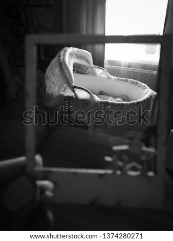 A rattan cradle with a baby next to the window. The black and white photo is taken through a square frame. A hand is holding the frame.