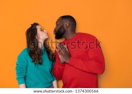 Profile photo of young beautiful people in love expressing warm feelings while kissing each other with closed eyes isolated over orange background. They are wearing casual clothes while posing for