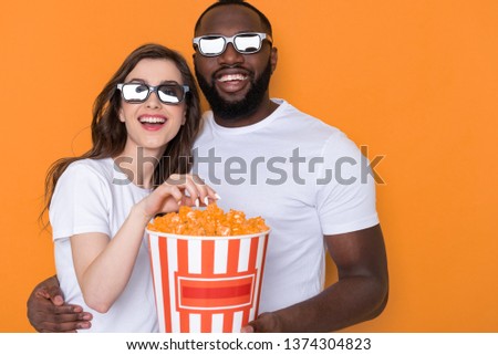 Close up of cheerful Caucasian girl wearing 3D glasses and looking at camera in studio. African guy keeping bucket with popcorn and watching a movie against orange background. Happy American man