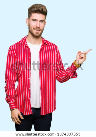 Young handsome man wearing red shirt with a big smile on face, pointing with hand and finger to the side looking at the camera.