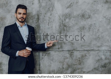 Waist up portrait of confident bearded young man in classic suit. He is standing with cup of coffee and pointing aside. Copy space in right side