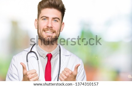 Young handsome doctor man wearing medical coat success sign doing positive gesture with hand, thumbs up smiling and happy. Looking at the camera with cheerful expression, winner gesture.