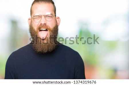 Young blond man wearing glasses and turtleneck sweater sticking tongue out happy with funny expression. Emotion concept.