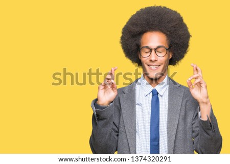 Young african american business man with afro hair wearing glasses smiling crossing fingers with hope and eyes closed. Luck and superstitious concept.