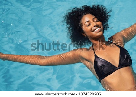 High angle view of beautiful african american girl with curly hair and bikini floating on back in swimming pool. Top view of smiling young woman carefree in pool with eyes closed relaxing in pool.