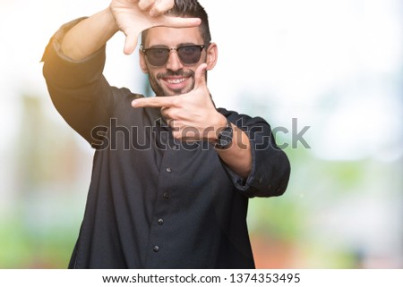 Young Christian priest wearing sunglasses over isolated background smiling making frame with hands and fingers with happy face. Creativity and photography concept.