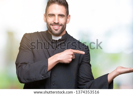Young Christian priest over isolated background amazed and smiling to the camera while presenting with hand and pointing with finger.