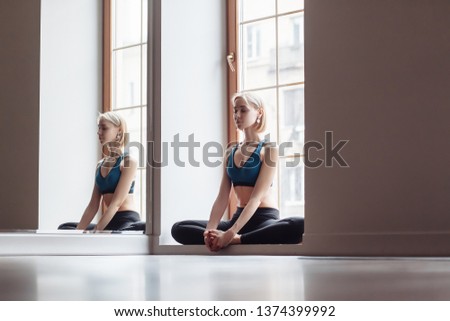 Charming female yoga instructor doing stretching exercise on the legs sitting on the mat on the background of a large window of her house. The concept of a healthy lifestyle and beautiful body
