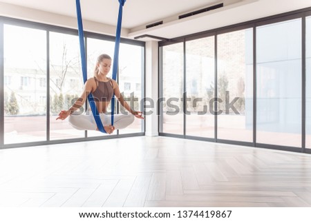 Beautiful young girl fitness instructor meditates sitting in lotus pose with eyes closed on hanging straps in a gym with large windows. Concept of advanced yoga lovers