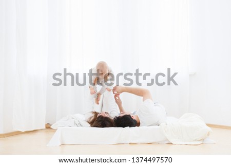 Portrait of young parents playing with little one-year old baby boy on bed indoors. Smiling baby raised hands up from pleasure. morning exercises for the baby with parents
