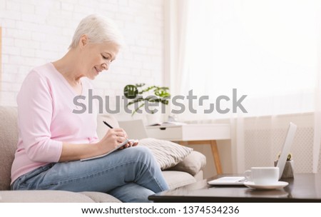 Advantages of modern technology. Senior woman rewriting recipe from laptop to notebook, searching in internet, empty space