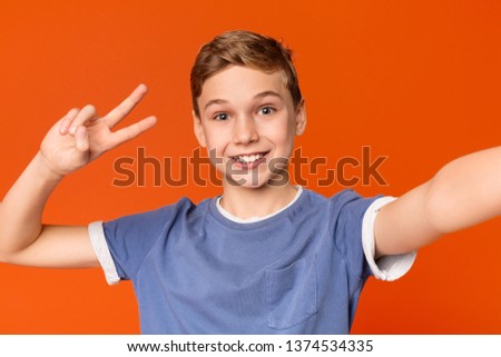 I love selfie! Adorable boy holding camera, making selfie and showing v-sign, standing against orange background
