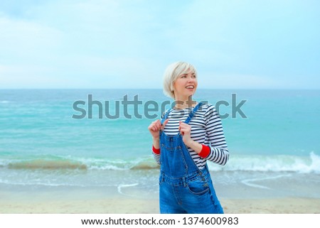 beautiful young blonde caucasian woman on vacation in striped blouse, sneakers and denim overall on the beach by the amazing blue sea background. Happy to be at the sea