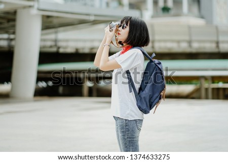 Portrait of fashionable young Asian woman photographer with vintage film camera and taking photo in the city. Making pictures in hipster style glasses and hat.