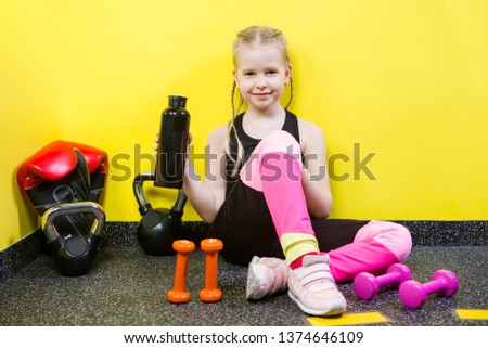 Theme sports and health children. Little Caucasian girl sits resting break floor gym holds hand bottle, drink water thirst. Athlete dumbbell equipments gymnastics bodybuilding background yellow wall.