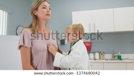 Cute little boy pretending to be a doctor with his mother while in pediatricians office using stethoscope. Child wearing oversized lab coat in exam room