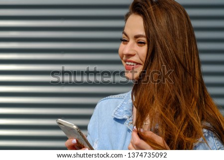A beautiful pretty modern young brunette with long hair in vintage jeans in a stylish denim jacket is resting near a modern metal building in the city in her hands . Fashion girl model.