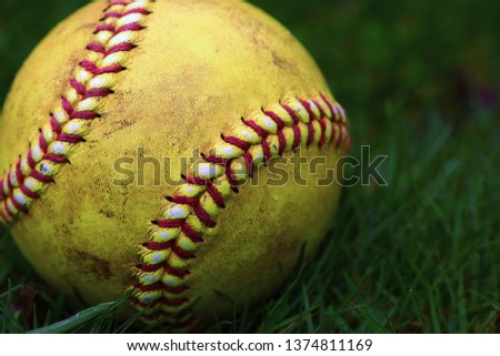 A yellow softball with red seams on grass.