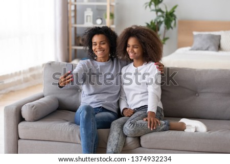 Smiling African American mother and teen daughter taking selfie for social network at home, sitting together on couch, using phone together, happy mum embracing child, posing for family photo