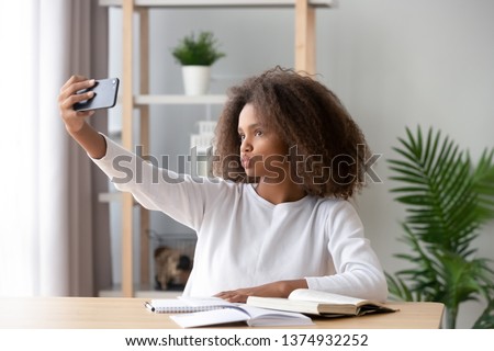 African American teenage girl taking selfie during homework preparation for social network blog, showing kiss, having fun with camera, posing for photo, distracted from school tasks, gadget addiction