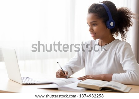 African American teen girl wearing headphones learning language online, using laptop, looking at screen, doing school tasks at home, writing notes, listening to lecture or music, distance education Royalty-Free Stock Photo #1374932258