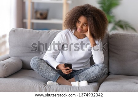 Unhappy thoughtful African American teenage girl sitting with phone, received unpleasant message, bad news, sitting on couch, feeling unhappy, teenage problem concept, first love and jealousy Royalty-Free Stock Photo #1374932342