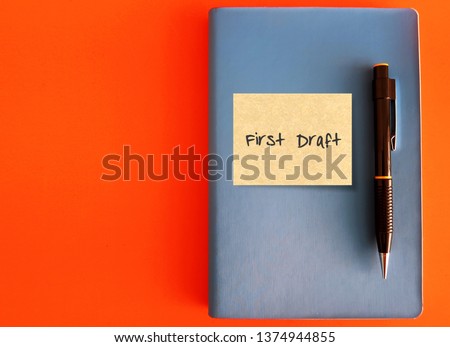 A blue notebook with sticky note written FIRST DRAFT and a black pen on orange background with copy space - concept of first draft book writing , screen writing, research or project planning