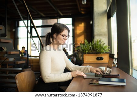 Smiling hipster girl in spectacles having online training course via laptop computer while sitting at workplace in coworking. Cheerful female blogger reading lifestyle article via notebook gadget 