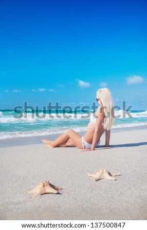 Cute blonde woman in white bikini and sunglasses on tropical sandy sea beach with seastars