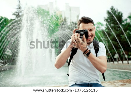 Outdoor summer lifestyle portrait of a handsome man having fun in city with camera in front of amazing fountain. Unshaven young boy using a retro photo camera
